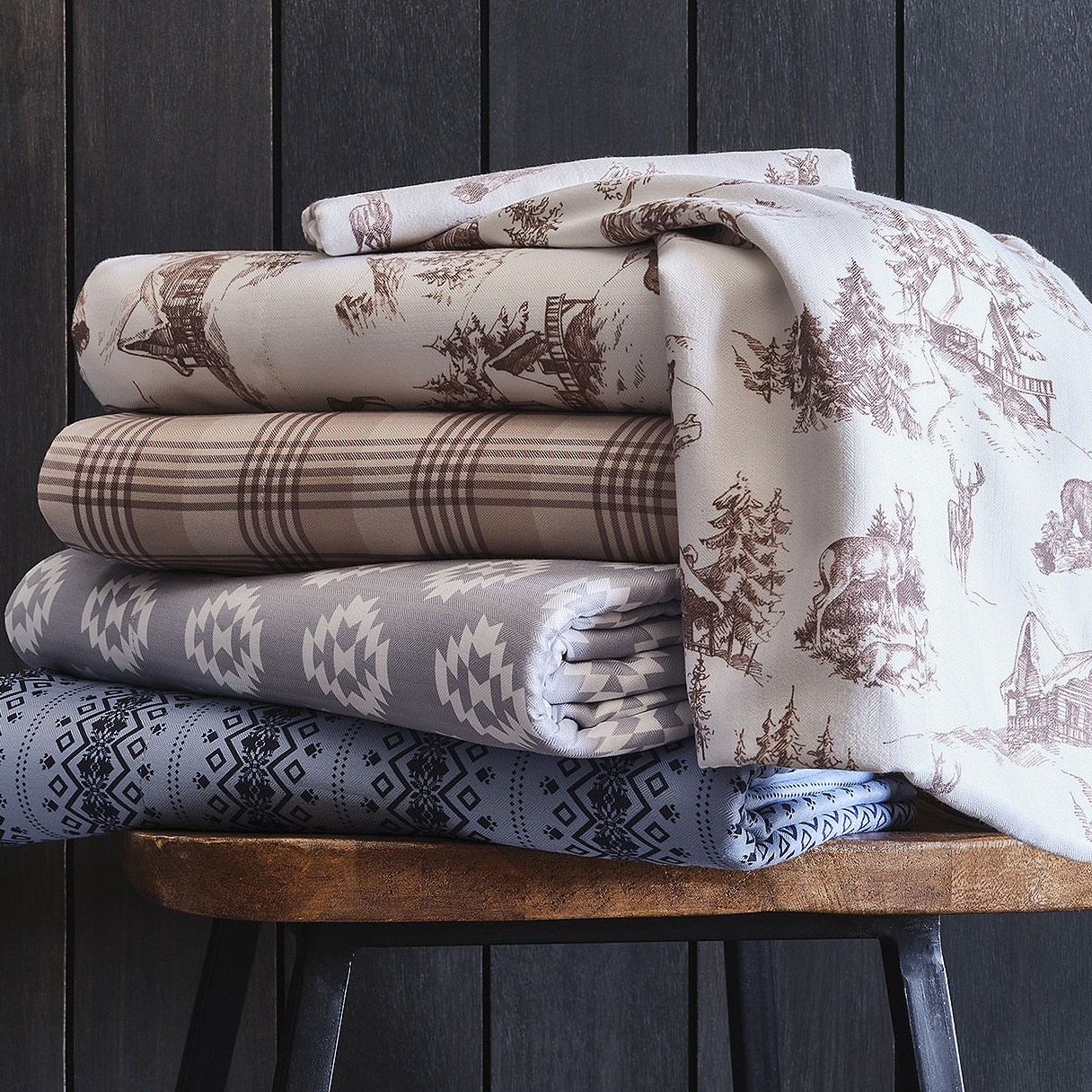 Stacked folded fabrics with varying patterns, including plaid and nature-inspired prints, on a wooden stool against a dark background.
