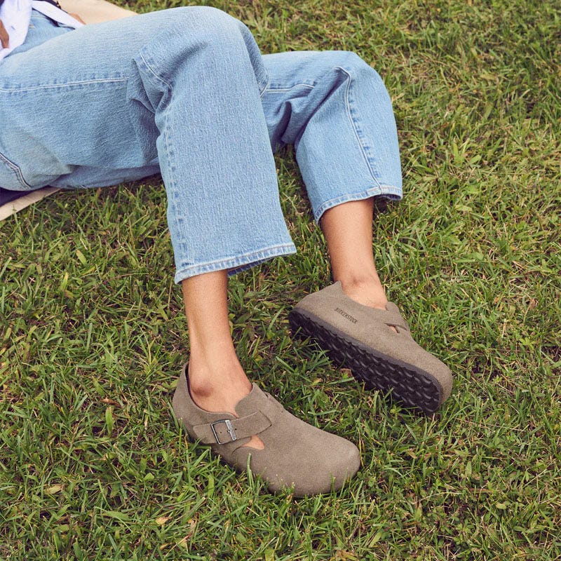 A person wearing light blue jeans and taupe-colored Birkenstock clogs with a buckle detail, sitting on grass.