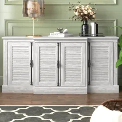 A gray, four-door sideboard with shutter-style doors and decorative metal handles is topped with a modern lamp, stacked books, and a vase of flowers, set against a light green paneled wall and a patterned rug on a wooden floor.