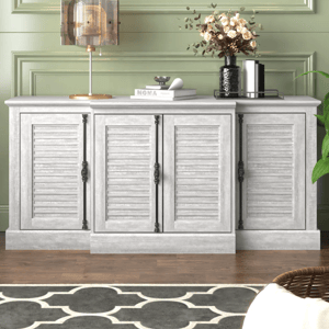 A gray, four-door sideboard with shutter-style doors and decorative metal handles is topped with a modern lamp, stacked books, and a vase of flowers, set against a light green paneled wall and a patterned rug on a wooden floor.