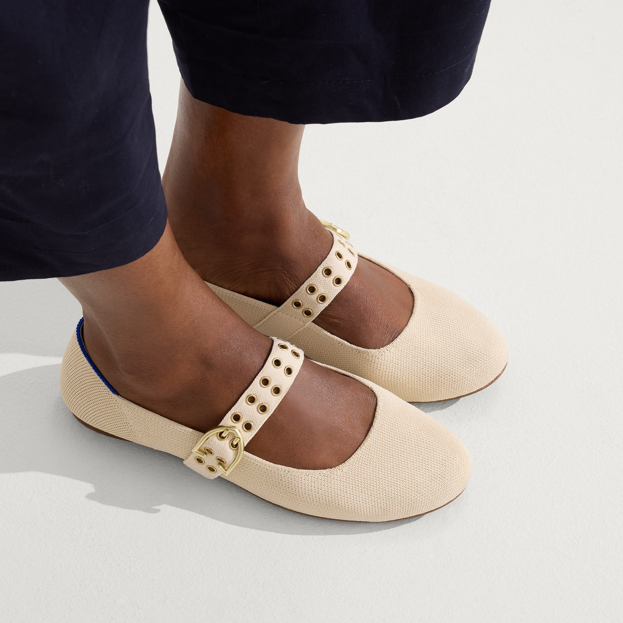 A pair of beige Mary Jane shoes with gold buckles and eyelet details is worn with dark pants.
