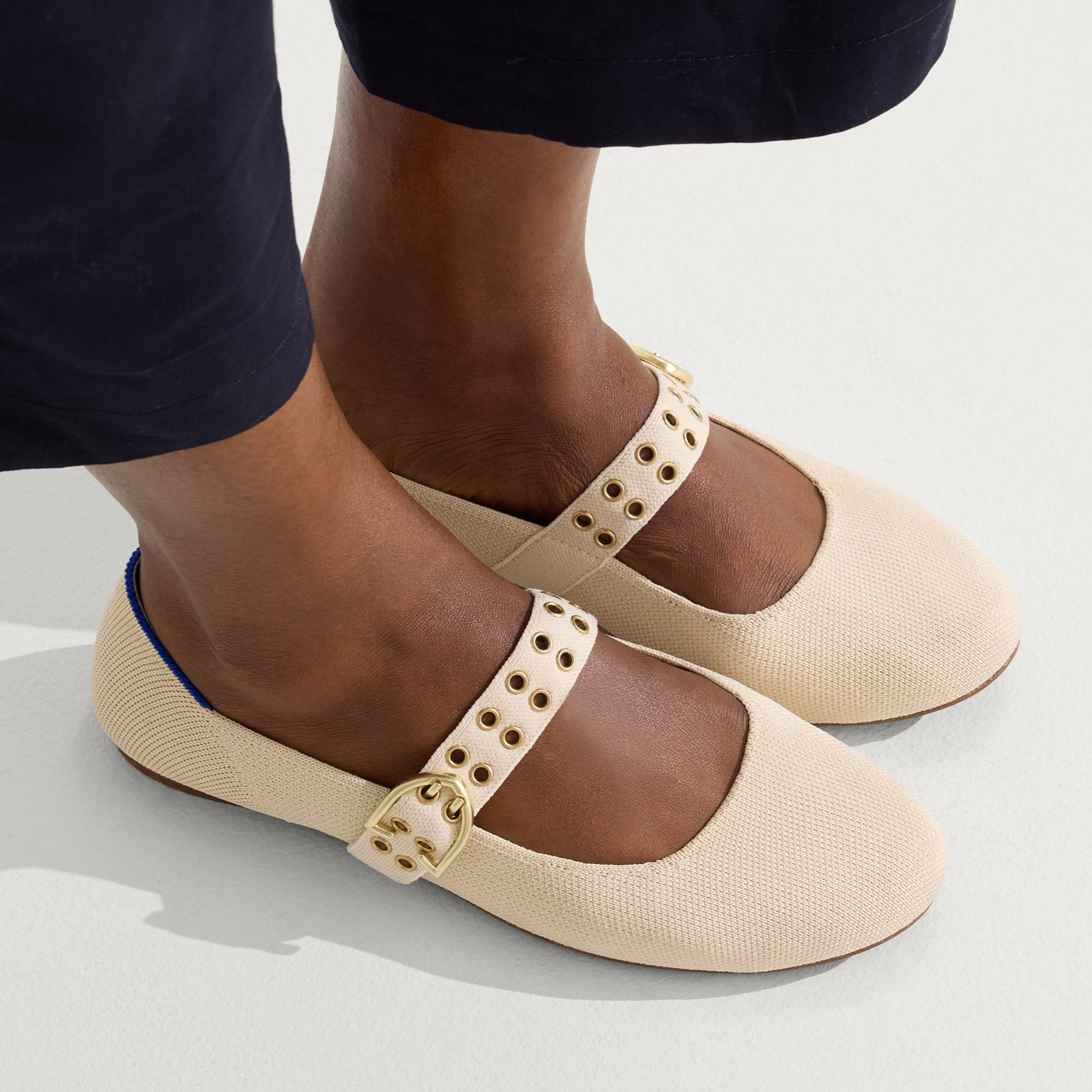 A pair of beige Mary Jane shoes with gold buckles and eyelet details is worn with dark pants.