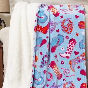 A cozy blanket features a fluffy white interior and a colorful exterior with patterns of cowboy boots, hats, and stars in pink, blue, and orange on a light blue background.
