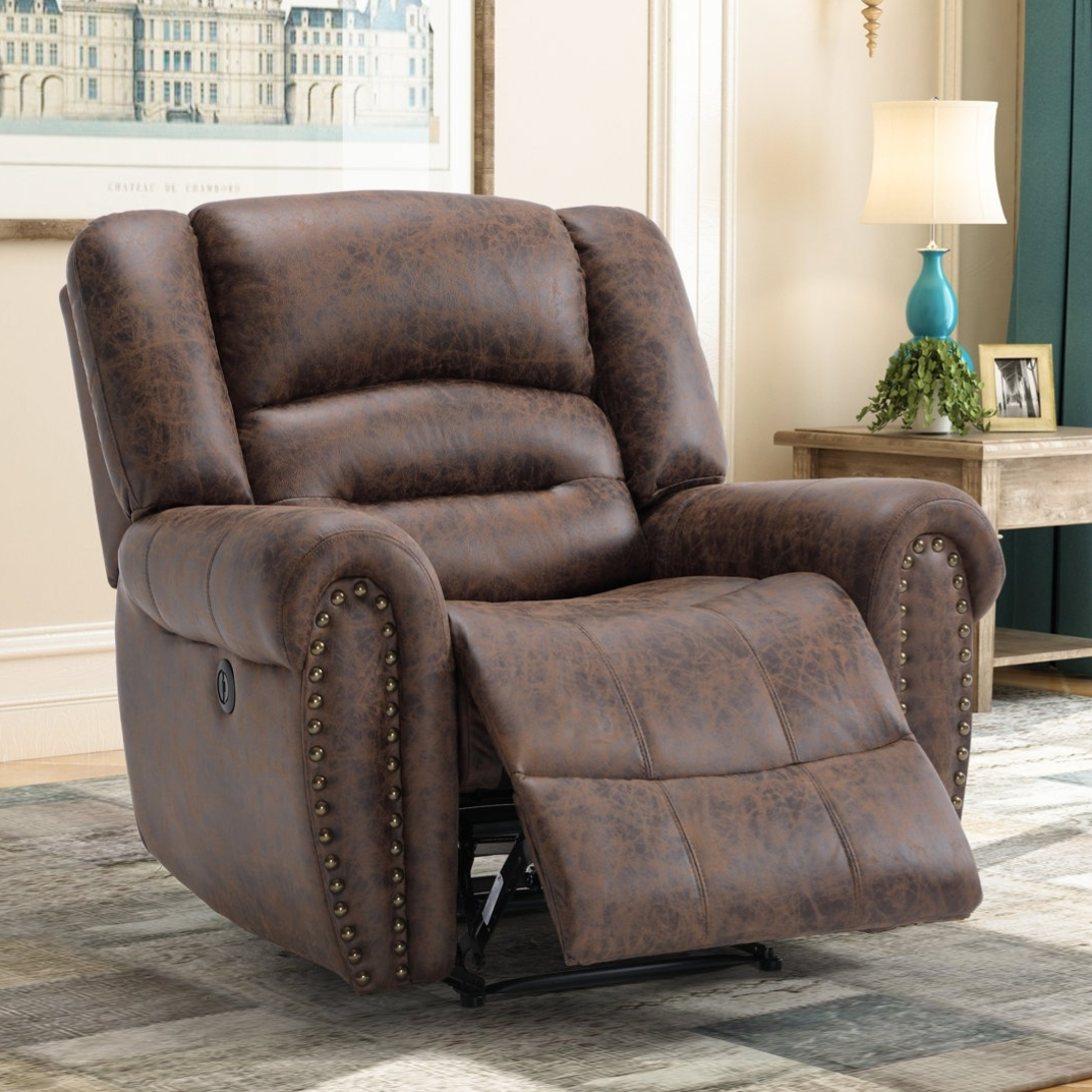 A brown leather recliner with nailhead trim is positioned in a living room setting, featuring a side table with a lamp and decorative items.