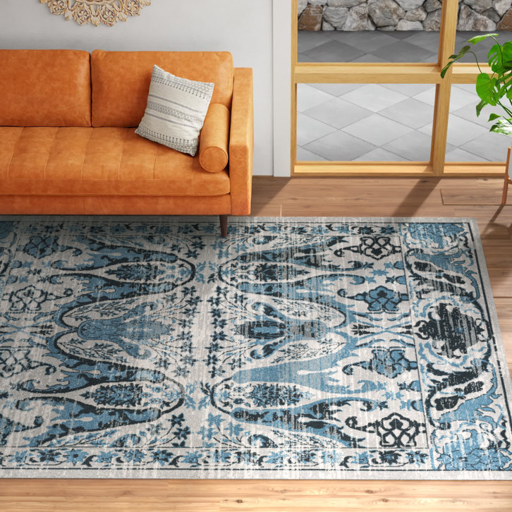 A large area rug with an intricate blue and black pattern against a light background, paired with an orange sofa and a decorative pillow, placed on wooden flooring adjacent to a large window and a potted plant.