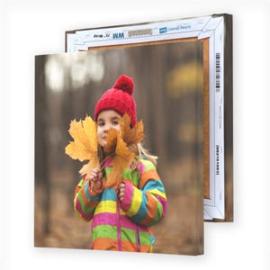 A canvas print shows a child in a colorful coat and red beanie holding large autumn leaves, with another identical canvas print partially visible behind it.