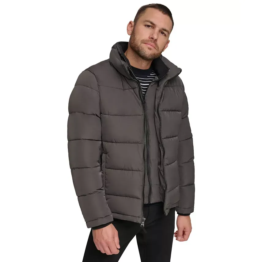 A man is wearing a dark gray, quilted puffer jacket with a stand-up collar. The jacket is paired with a striped shirt underneath and black pants.