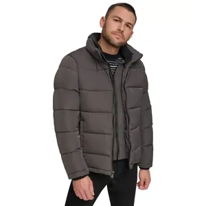 A man is wearing a dark gray, quilted puffer jacket with a stand-up collar. The jacket is paired with a striped shirt underneath and black pants.