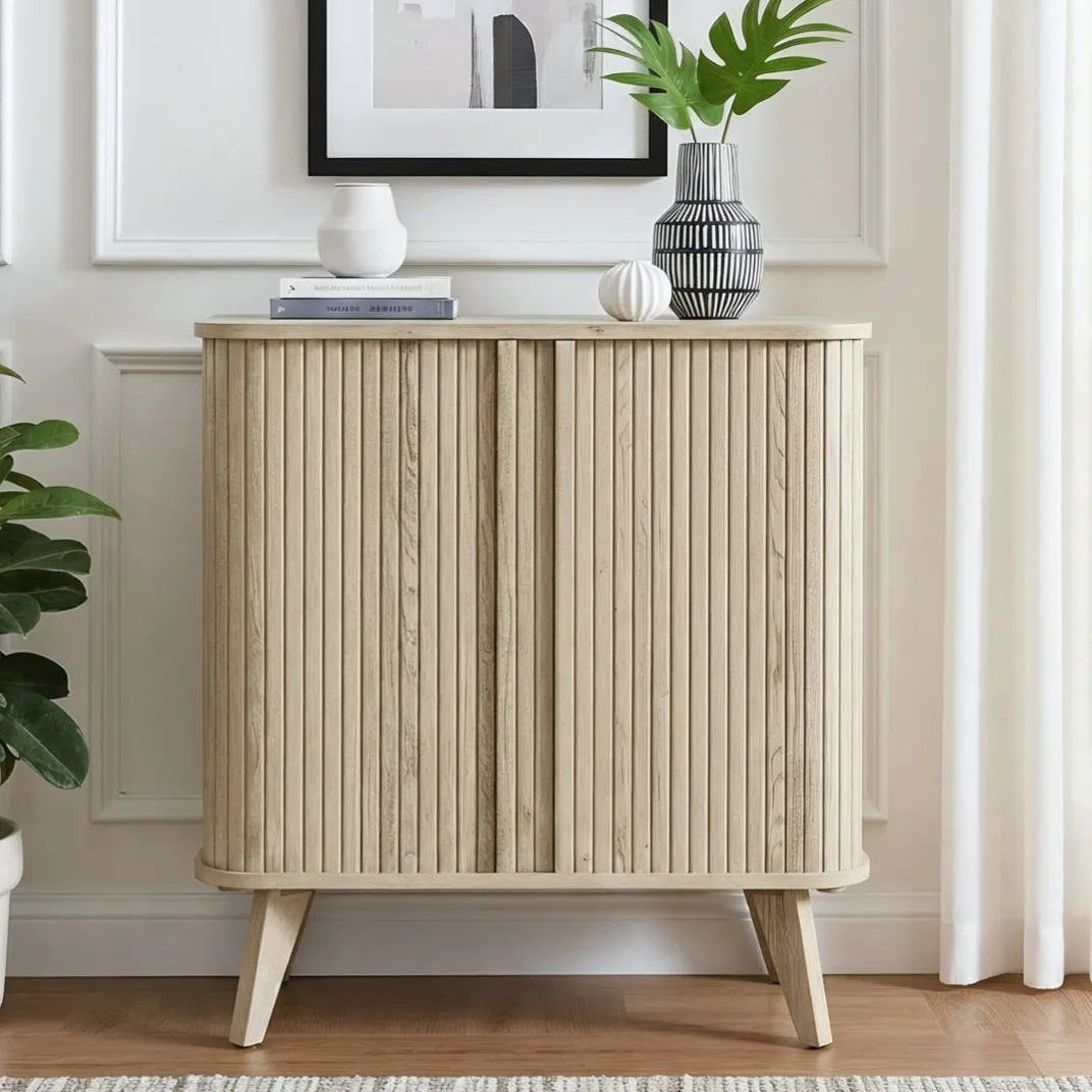 A light wooden cabinet with vertical grooves, featuring angled legs, is topped with decorative vases and books, accompanied by a potted plant on the side, all set against a white paneled wall with an abstract framed artwork.