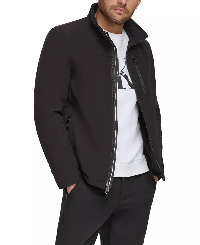 A man is wearing a black zip-up jacket with a stand collar over a white shirt and black pants, featuring zippered pockets.