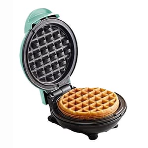 A round, open mini waffle maker with a light green exterior and non-stick black grid plates, featuring a cooked waffle inside.