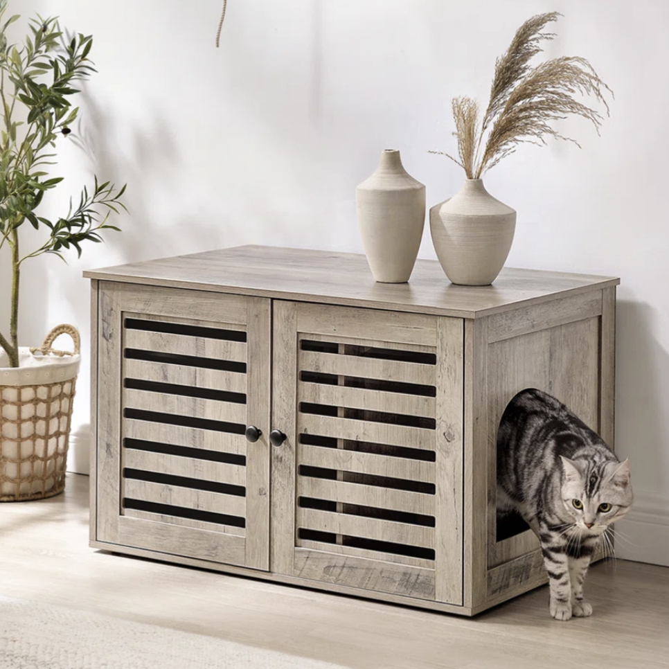A wooden pet furniture with louvered doors and a side entrance is shown, accompanied by decorative vases on top. A cat is emerging from the entrance. The setting includes a leafy plant in a basket and a cozy beige sofa.