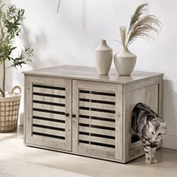 A wooden pet furniture with louvered doors and a side entrance is shown, accompanied by decorative vases on top. A cat is emerging from the entrance. The setting includes a leafy plant in a basket and a cozy beige sofa.