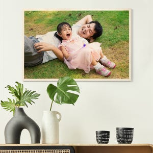 A custom wood print displaying a joyful moment with a parent and child laughing on grass, housed in a minimalist room with decorative vases and plants.