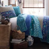 Blue and teal patchwork quilt and pillows on a bed, accompanied by rustic decor including a wicker basket with books, decorative spheres, a ceramic vase, and wooden chest.