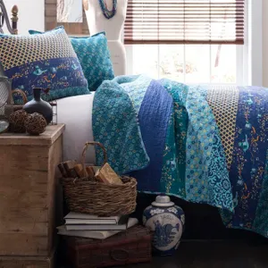 Blue and teal patchwork quilt and pillows on a bed, accompanied by rustic decor including a wicker basket with books, decorative spheres, a ceramic vase, and wooden chest.