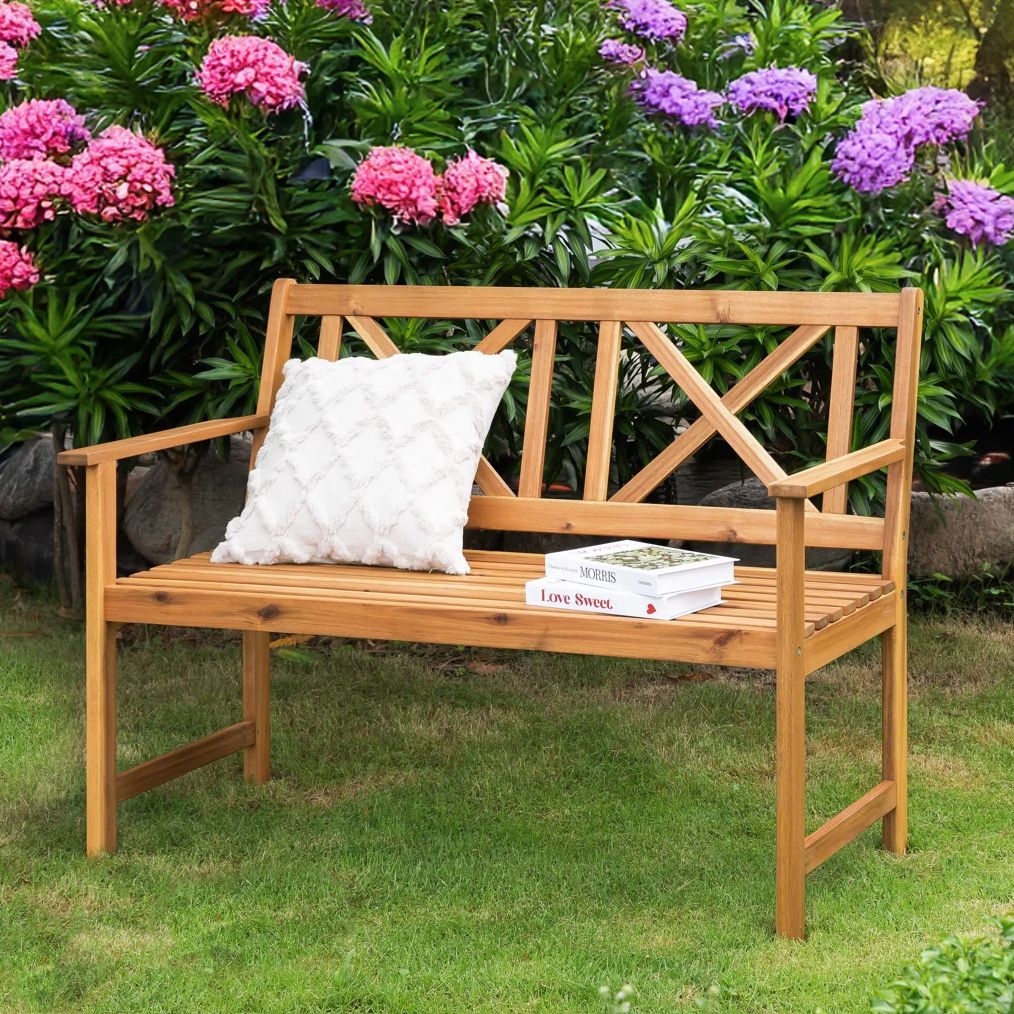 Wooden bench with crisscross backrest, white textured pillow, and two books titled \“MORRIS\“ and \“Love Sweet\“ on grass, surrounded by vibrant pink and purple flowers.