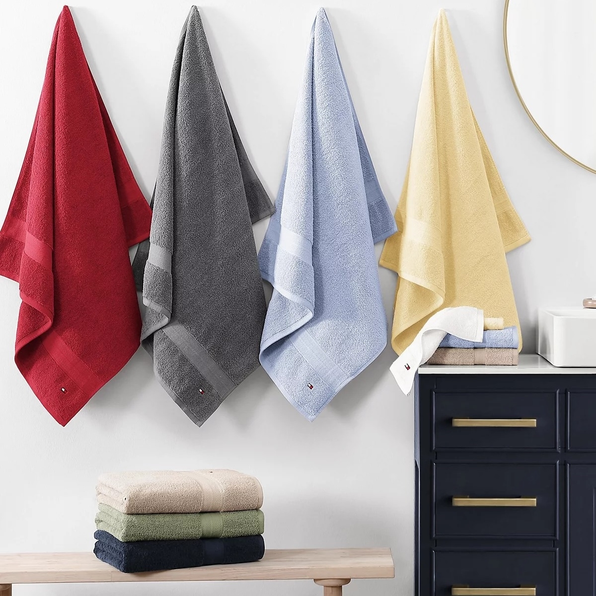 Four hanging towels in red, gray, blue, and yellow are displayed alongside folded towels in beige, green, and navy on a wooden bench near a dark navy drawer with gold handles.