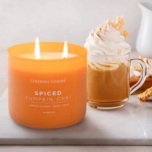 Orange candle labeled \“Colonial Candle Spiced Pumpkin Chai\“ next to a glass mug filled with a pumpkin-themed beverage topped with whipped cream and spices.