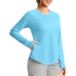 A woman is wearing a light blue long-sleeve shirt paired with gray shorts. The shirt has thumbholes and a relaxed fit, suitable for casual or athletic use.