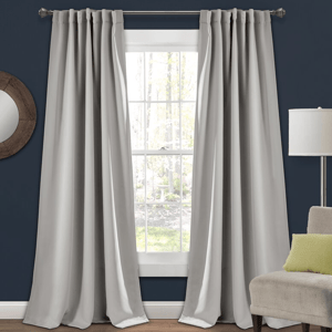 Set of 2 light gray blackout curtain panels hang on a rod in front of a window, blocking light and adding privacy, with a smooth fabric finish.