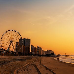 A ferris wheel by the beach at sunset with a pier extending into the water and high-rise buildings in the background.