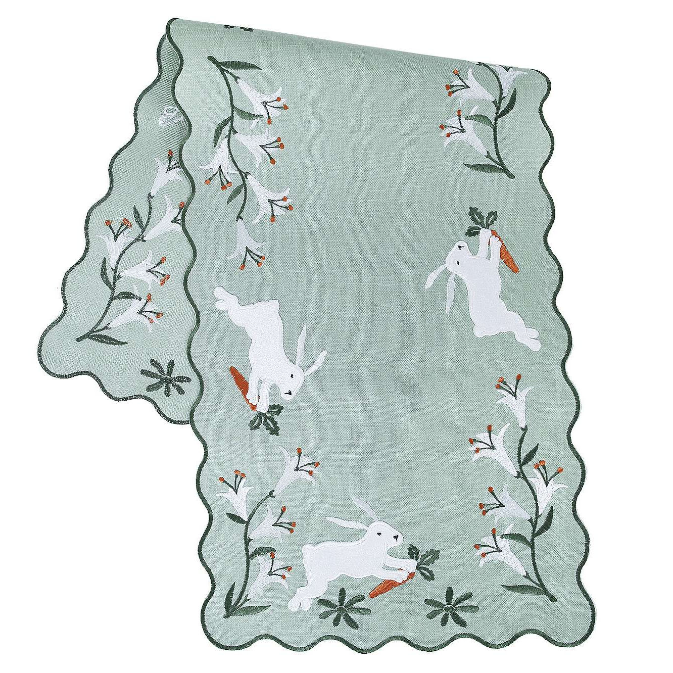 Green table runner with scalloped edges, featuring embroidered white bunnies holding carrots and surrounded by floral designs with orange and white flowers.