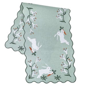 Green table runner with scalloped edges, featuring embroidered white bunnies holding carrots and surrounded by floral designs with orange and white flowers.