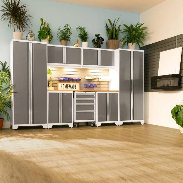 7-piece NewAge garage system features sleek gray cabinets with white frames, high and low storage units, a workbench, drawers on wheels, and overhead cabinets, all surrounded by potted plants on top for a tidy, organized workspace.