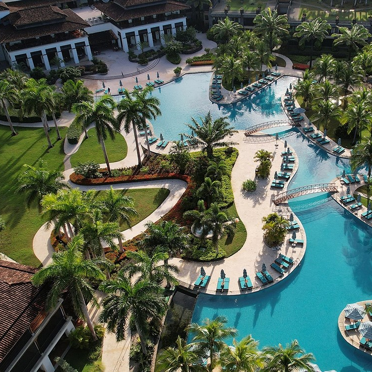 Aerial view of a resort featuring lush greenery, multiple winding pools, lounge chairs under umbrellas, and elegant landscaping with palm trees.