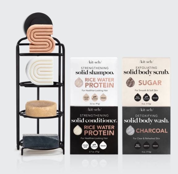 A black three-tier metal shower caddy holds four solid personal care items; shampoo, conditioner, body wash, and body scrub, all labeled with their respective ingredients.