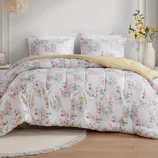 A floral-patterned bedding set with a comforter and pillows, featuring pastel colors on a white background, complemented by a yellow throw. A round, fringed rug lies underneath.