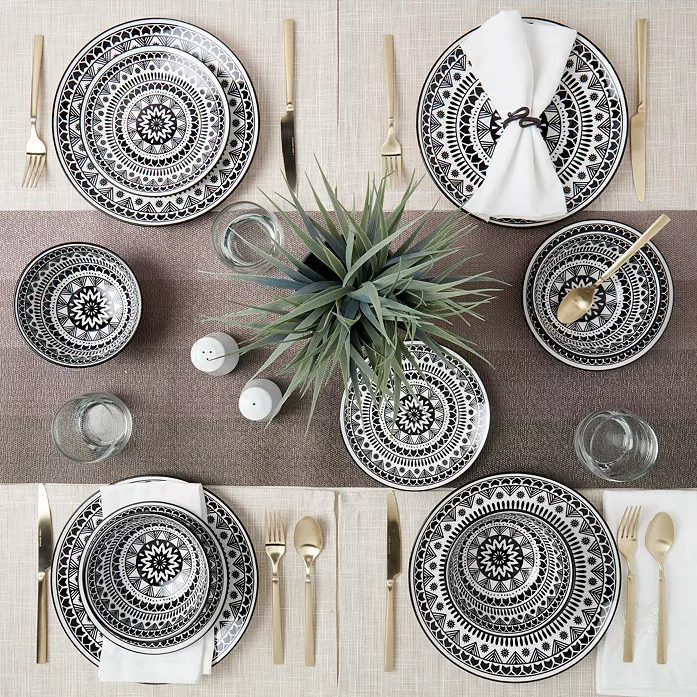 This is a set of decorative black and white patterned dinnerware with matching gold flatware, displayed on a taupe woven tablecloth.