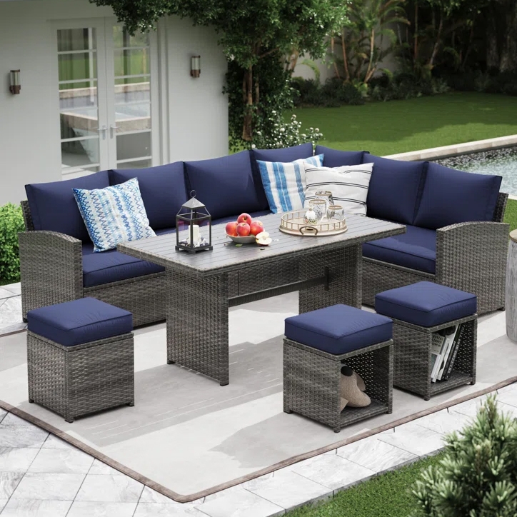 Outdoor patio set featuring a sectional sofa with navy cushions, a rectangular table, and matching stools on a tiled patio. Decorative pillows, a lantern, and a tray with snacks are on the table, set in a garden with a pool in the background.