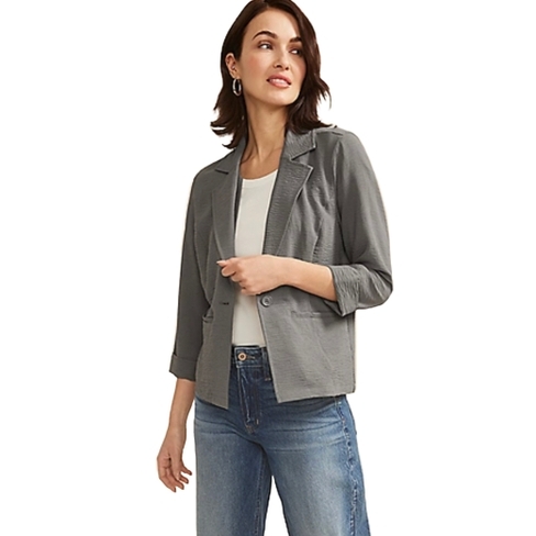 A woman is wearing a gray blazer over a white top paired with blue jeans. The blazer has three-quarter sleeves and a relaxed fit, accompanied by hoop earrings.