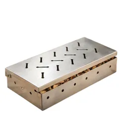 A stainless steel smoker box filled with wood chips is on a table beside grilled meat, tomatoes, and corn.