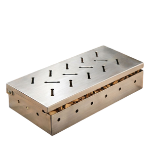 A stainless steel smoker box filled with wood chips is on a table beside grilled meat, tomatoes, and corn.