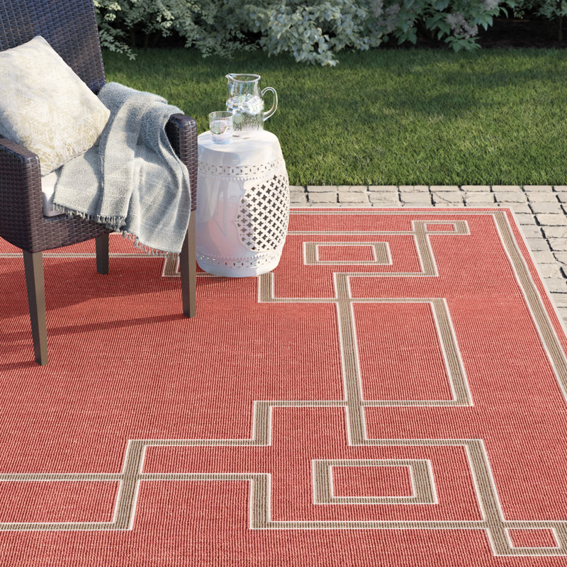 Outdoor setup featuring a wicker chair with a pillow and throw, a white ceramic side table, and a red geometric-patterned rug on a paved patio.