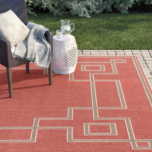 Outdoor setup featuring a wicker chair with a pillow and throw, a white ceramic side table, and a red geometric-patterned rug on a paved patio.