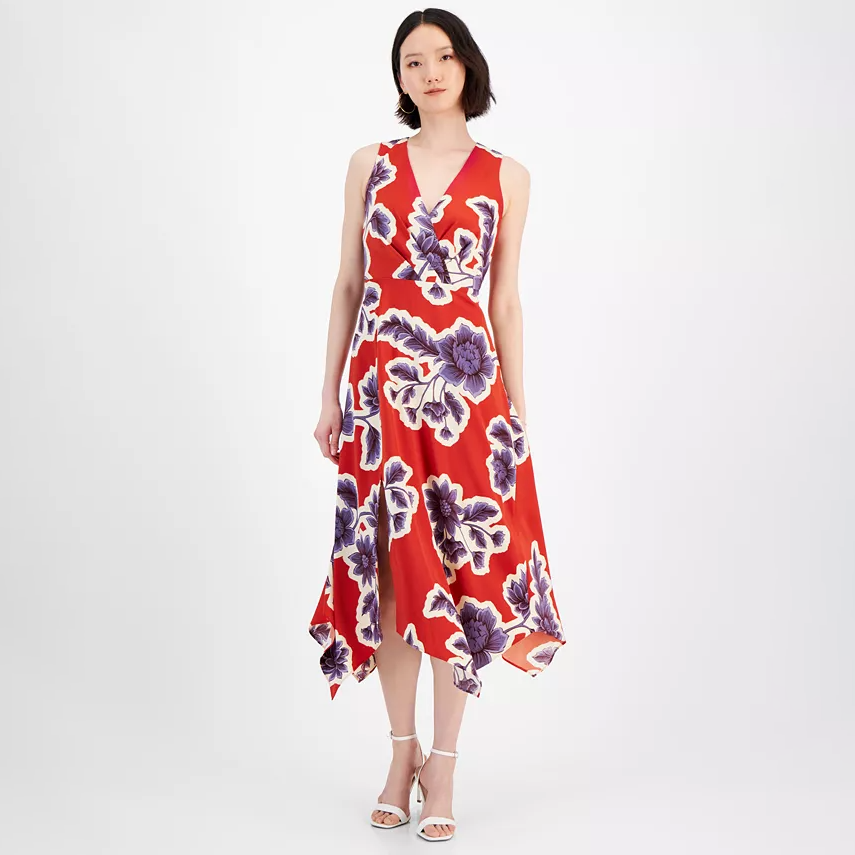 A red midi dress with a v-neckline featuring a large purple and white floral print, asymmetrical hem, and sleeveless design. The model is wearing white strappy heels.