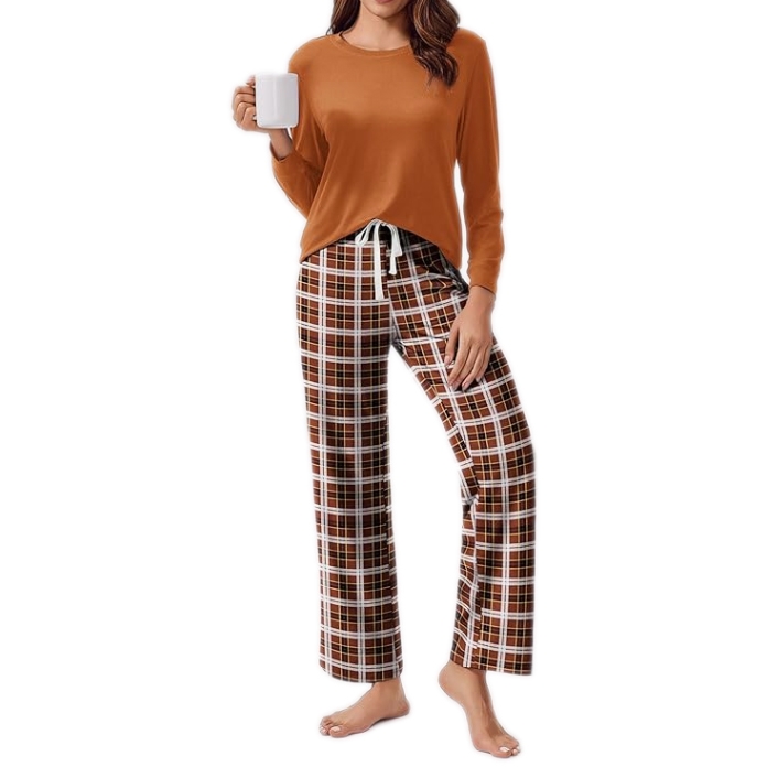 A woman is wearing a two-piece pajama set with a solid orange long-sleeve top and flared plaid pants in brown, white, and red. She is holding a white mug and standing barefoot.