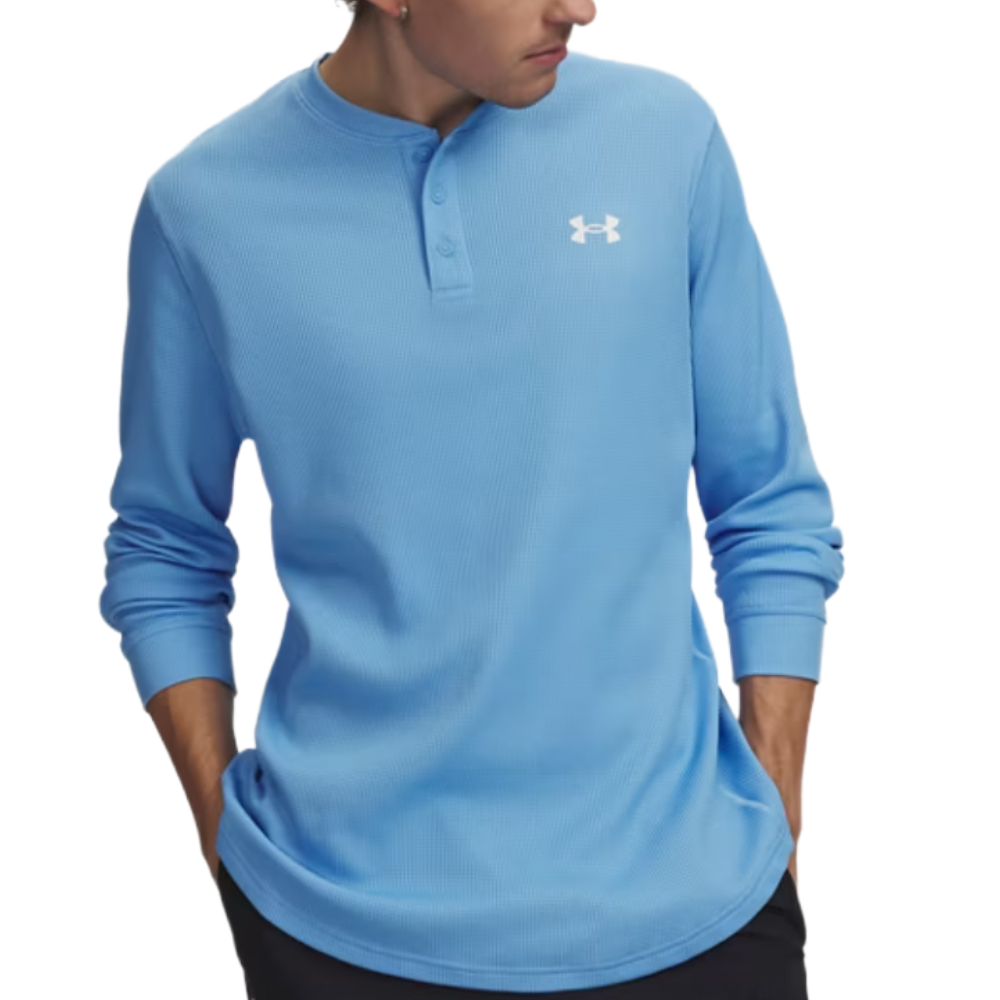 A person wearing a light blue Under Armour Henley shirt with a logo on the chest and black pants, hands in pockets.