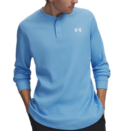 A person wearing a light blue Under Armour Henley shirt with a logo on the chest and black pants, hands in pockets.