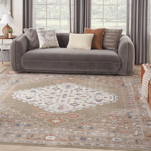 A modern living room features a gray sofa adorned with four decorative pillows in various patterns and colors. The floor showcases a large, intricately designed area rug with a central geometric motif and muted tones.