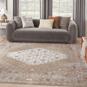 A modern living room features a gray sofa adorned with four decorative pillows in various patterns and colors. The floor showcases a large, intricately designed area rug with a central geometric motif and muted tones.