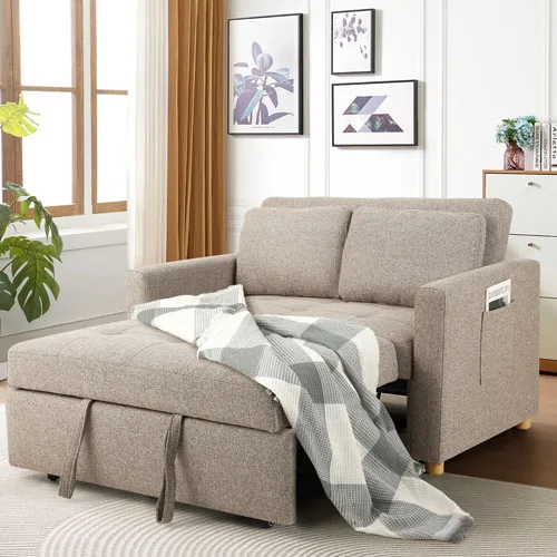 A stylish beige sofa bed with a checkered blanket draped over it, featuring storage space and side pockets. Wall art and indoor plants enhance the cozy living room setting.