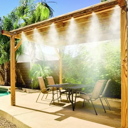 A pergola with an integrated outdoor misting system provides cooling over a patio area with a table and chairs, set beside a pool and surrounded by lush greenery.