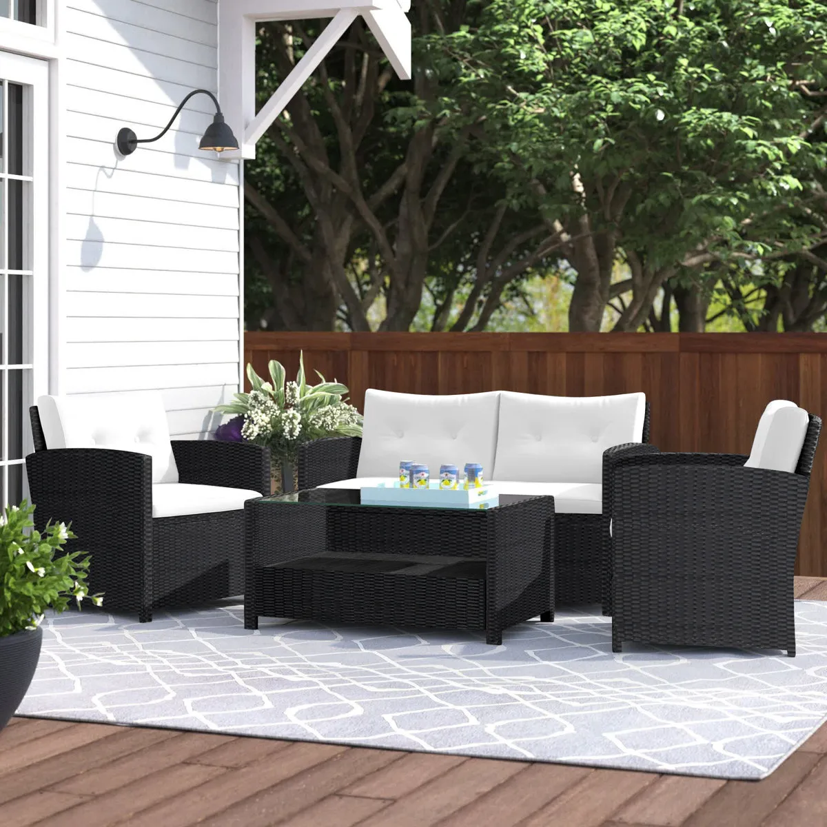 Outdoor patio set with a black wicker design featuring a white cushioned loveseat, two armchairs, and a glass-top coffee table on a patterned blue and white rug, set on a wooden deck near a white house and surrounded by greenery.