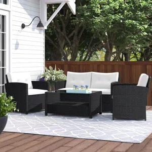 Outdoor patio set with a black wicker design featuring a white cushioned loveseat, two armchairs, and a glass-top coffee table on a patterned blue and white rug, set on a wooden deck near a white house and surrounded by greenery.