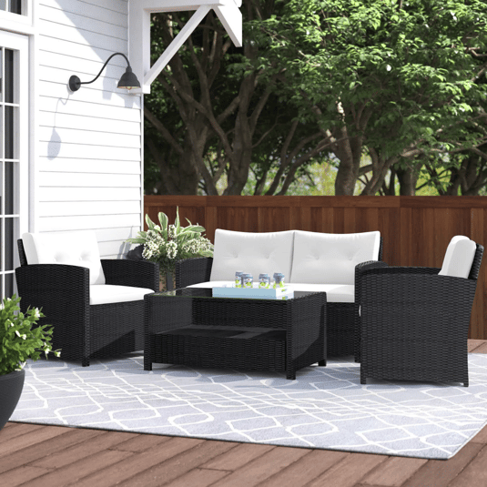 Outdoor patio set with a black wicker design featuring a white cushioned loveseat, two armchairs, and a glass-top coffee table on a patterned blue and white rug, set on a wooden deck near a white house and surrounded by greenery.